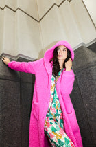 A Lady Wearing Maisie Sherpa Long Jacket In Holly Jolly Pink with the hood up, standing against a building wall